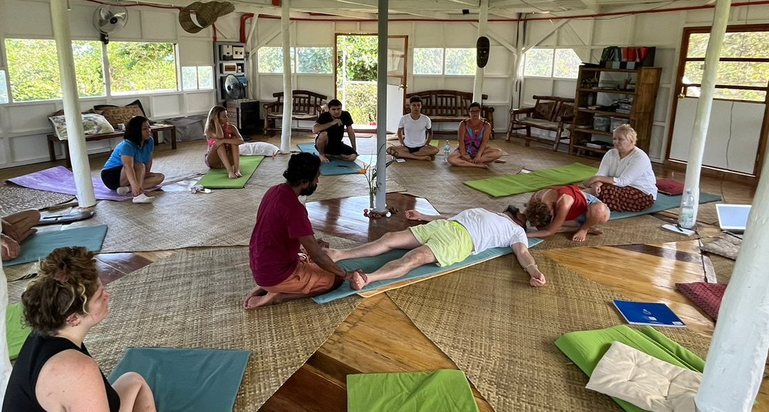 A group sitting in a circle on mats in an open wooden space, with one person lying down at the centre during a session