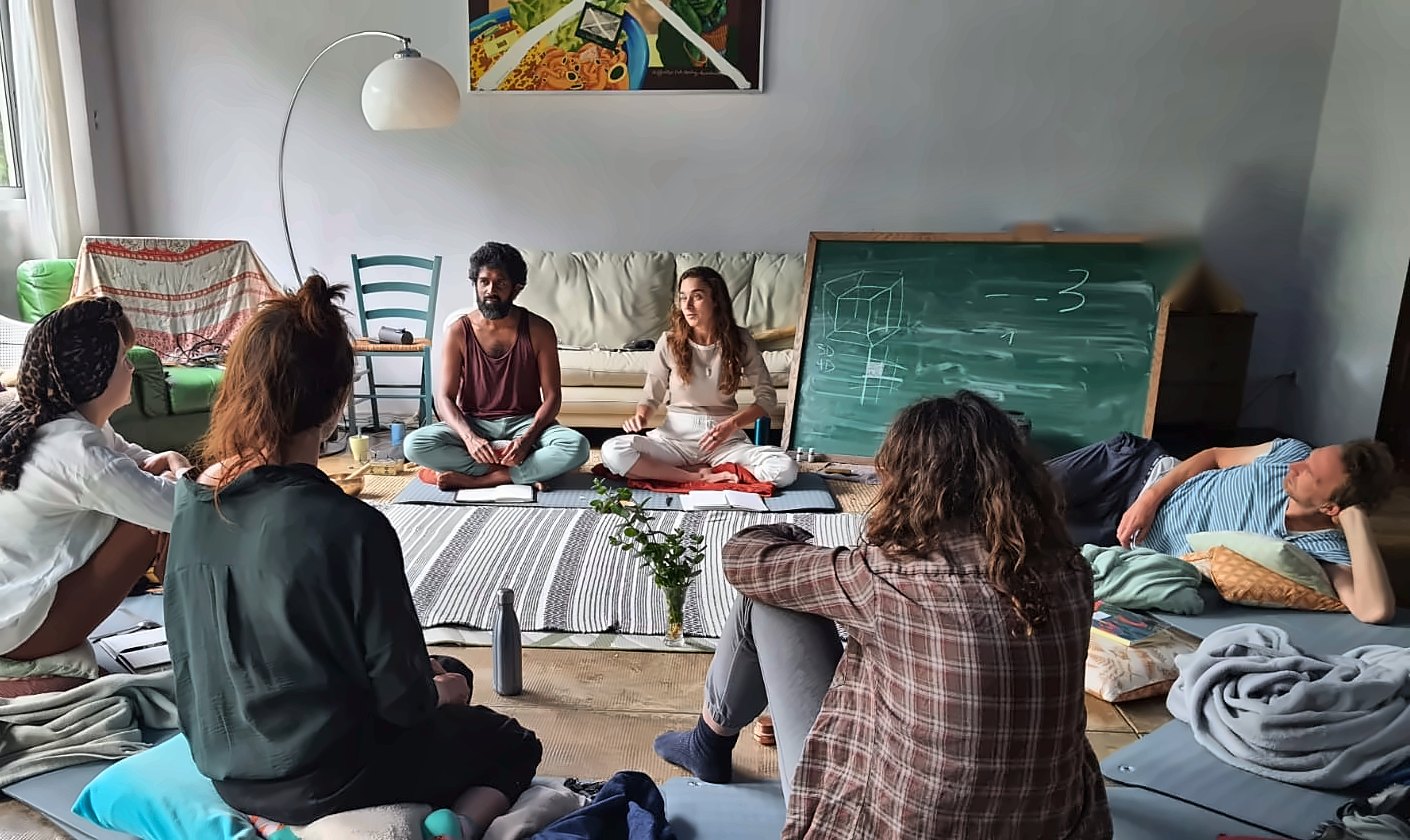 Ahilan und Fanny unterrichten eine kleine Gruppe, die eng beieinander sitzt, mit einer handgezeichneten Skizze auf einer Tafel im Hintergrund