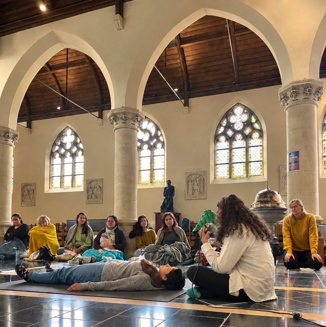 Teilnehmende versammelt in einer Steinkirche mit Bogenfenstern: Eine Person ruht auf einer Matte, während ein Facilitator zur Gruppe spricht