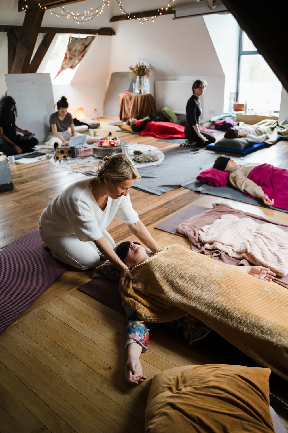 Eine Facilitatorin kniet neben einer Teilnehmerin, die mit Augenmaske unter einer Decke liegt, in einem Raum, in dem weitere Teilnehmende auf Matten ruhen