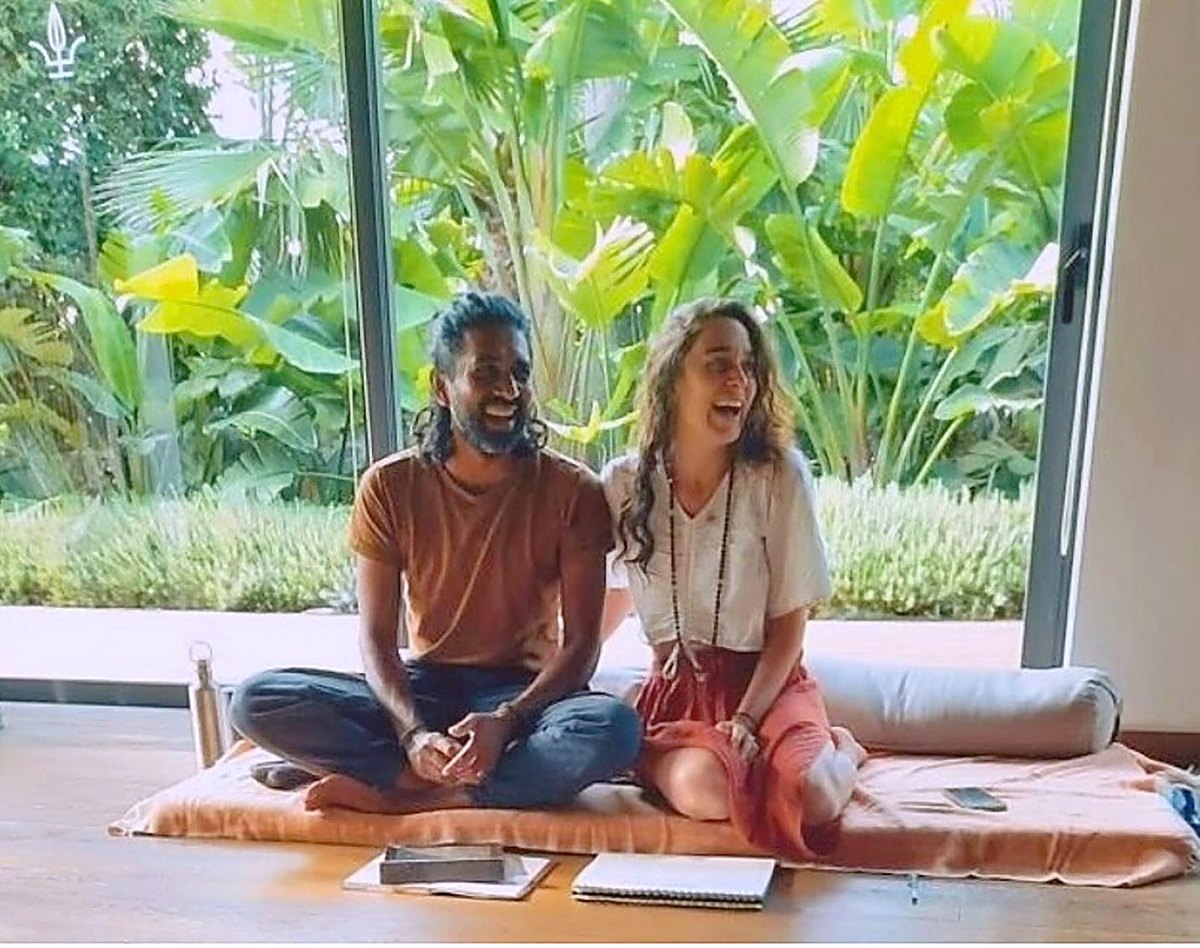 Ahilan und Fanny sitzen im Schneidersitz auf einer Matte vor einem Garten und lachen zusammen